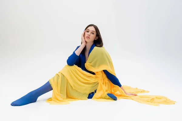 Patriotische ukrainische junge Frau in blau-gelbem Outfit mit Strumpfhosen sitzt, während sie auf grau posiert — Stockfoto