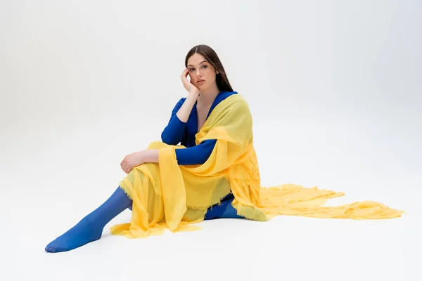 Dreamy ukrainian young woman in blue and yellow outfit with tights sitting while posing on grey — Stock Photo