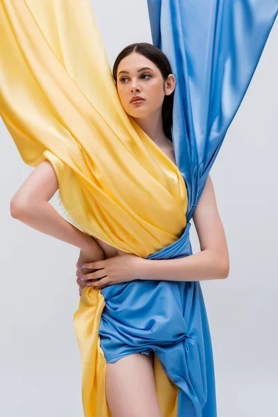 Mujer joven ucraniana en vestido azul y amarillo posando con la mano en la cadera aislada en gris - foto de stock