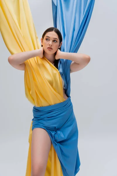 Брюнетка украинская женщина в синем и желтом платье смотрит в сторону и позирует изолированно на сером — стоковое фото