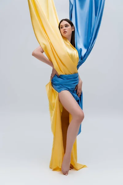 Full length of brunette and young ukrainian woman in blue and yellow dress posing on grey — Stock Photo