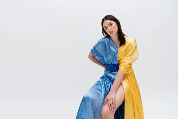 Чувственная молодая украинка в синем и желтом платье, позирующая рукой на бедре, сидя изолированная на сером — стоковое фото