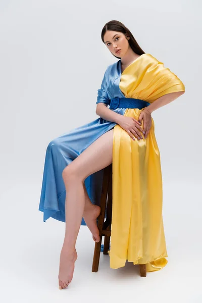 Barefoot ukrainian woman in blue and yellow dress posing while sitting on wooden chair on grey — Fotografia de Stock