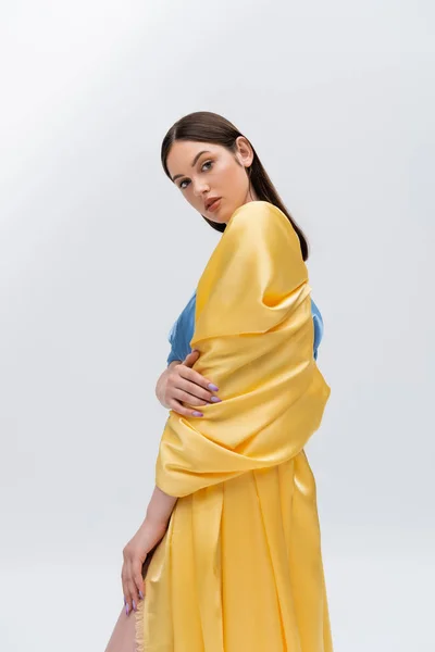 Чувственная молодая украинская модель в синем и желтом платье, позирующая при взгляде на камеру, изолированную на сером — стоковое фото