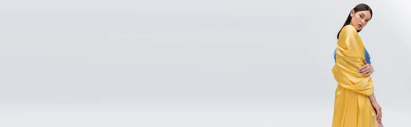 Чувственная молодая украинская модель в синем и желтом платье позирует при взгляде на камеру, изолированную на сером, баннер — стоковое фото