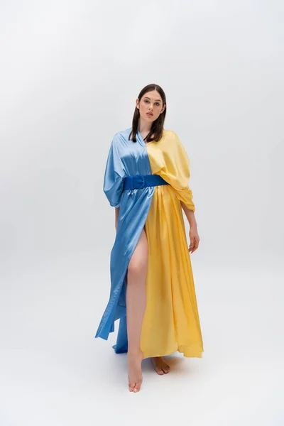 Full length of barefoot young ukrainian woman in blue and yellow dress posing on grey — Stockfoto