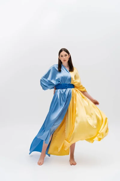 Full length of barefoot young ukrainian woman in blue and yellow dress posing with hand on hip on grey — Stock Photo