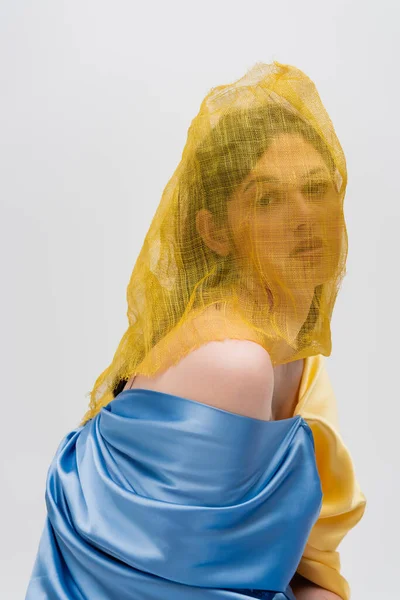 Mujer joven ucraniana con la cara cubierta de cortinas amarillas mirando a la cámara aislada en gris - foto de stock