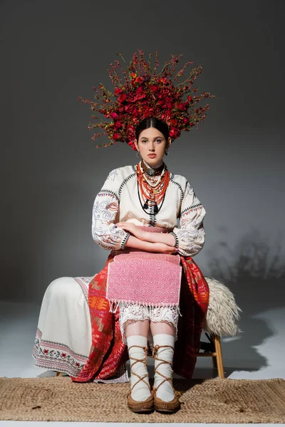 Full length of young ukrainian woman in traditional clothing and floral red wreath sitting on bench on grey — Photo de stock