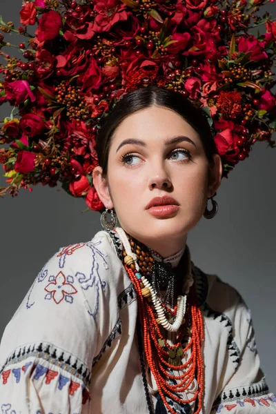 Portrait of ukrainian woman in traditional clothing and floral red wreath looking away isolated on grey — Stockfoto