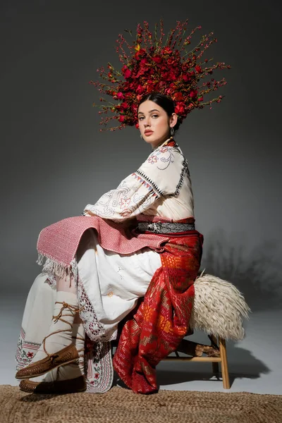 Полная длина красивой украинской женщины в традиционной одежде и красном венке с цветами и ягодами, сидящими на сером — стоковое фото