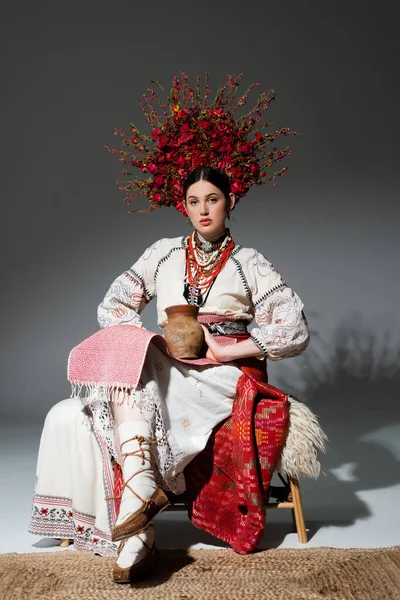 Pleine longueur de jeune femme ukrainienne en couronne rouge avec des fleurs et des baies tenant pot d'argile sur gris foncé — Photo de stock