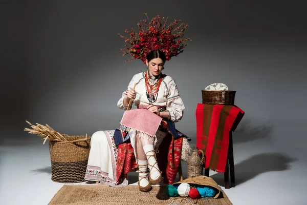 Full length of ukrainian woman in traditional clothes and red floral wreath holding knitting needles on dark grey — Fotografia de Stock