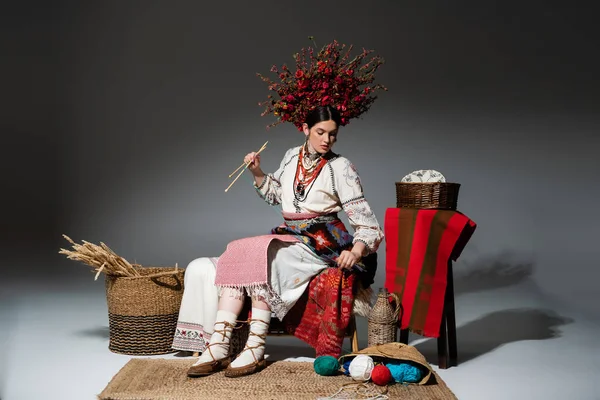 Full length of young ukrainian woman in traditional clothes and red wreath with flowers holding knitting needles on dark grey — Stockfoto