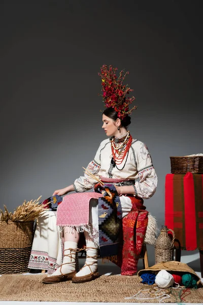 Comprimento total da mulher ucraniana em roupas tradicionais e grinalda vermelha com flores segurando agulhas de tricô no cinza escuro — Fotografia de Stock