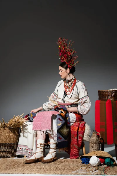 Full length of ukrainian woman in traditional clothes and red wreath with flowers holding knitting needles on dark grey - foto de stock