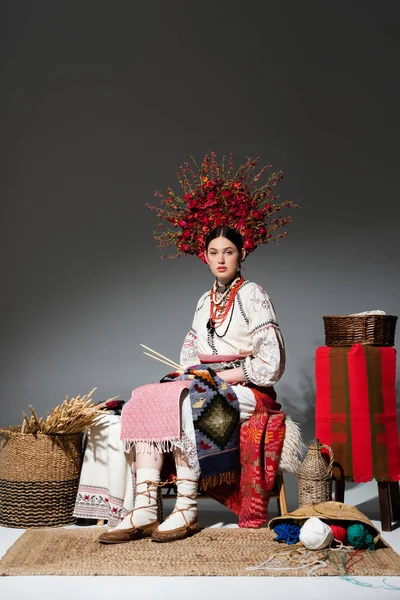 Pleine longueur de femme ukrainienne brune en vêtements traditionnels et couronne de fleurs tenant aiguilles à tricoter sur gris foncé — Photo de stock