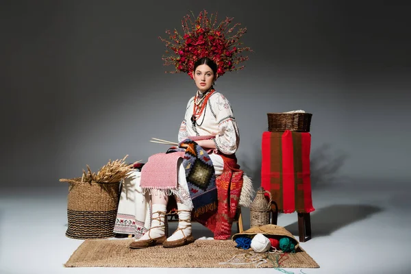 Full length of young ukrainian woman in traditional clothes and red floral wreath holding knitting needles on dark grey — Stockfoto