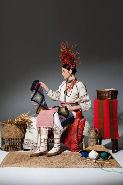 Full length of young ukrainian woman in traditional clothes and red floral wreath holding knitted scarf on dark grey - foto de stock