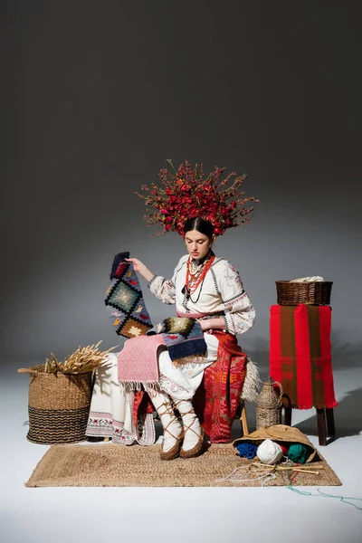 Full length of brunette ukrainian woman in traditional clothes and red floral wreath holding knitted scarf on dark grey — Stock Photo