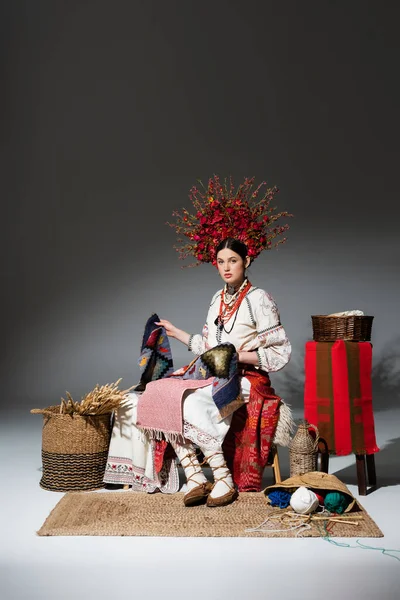 Full length of young ukrainian woman in traditional clothes and floral wreath with berries holding knitted scarf on dark grey — Fotografia de Stock