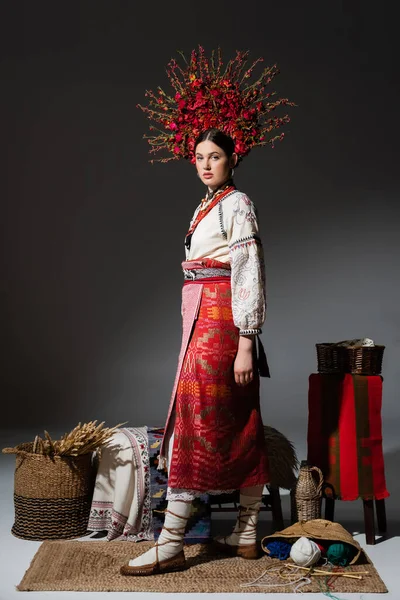 Full length of ukrainian woman in traditional red floral wreath standing near yarn balls and wheat on dark grey - foto de stock