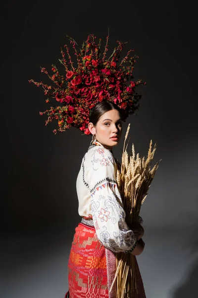 Bruna e giovane donna ucraina in ghirlanda floreale con bacche che tengono il grano sul nero — Foto stock