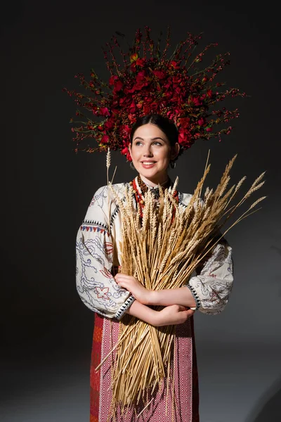 Mujer ucraniana feliz y joven en corona floral con bayas rojas sosteniendo espiguillas de trigo en negro - foto de stock