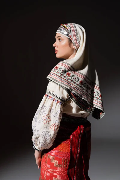 Vue latérale de la femme ukrainienne en chemise traditionnelle avec ornement et coiffure regardant loin sur noir — Photo de stock