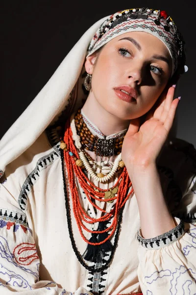 Portrait of ukrainian woman in traditional shirt with red ornament and headwear looking away isolated on black - foto de stock