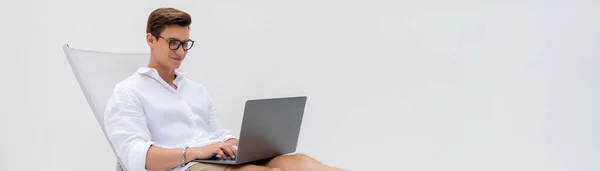 Щасливий чоловік в окулярах використовує ноутбук у кріслі палуби ізольований на білому, банер — стокове фото