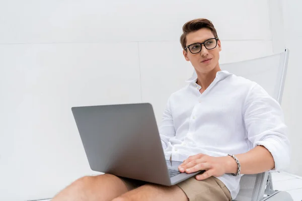 Smiling freelancer sitting in deck chair with laptop and looking away — Stockfoto