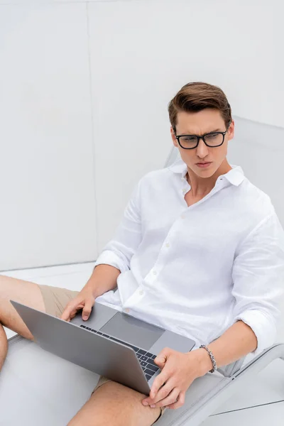 Thoughtful man in eyeglasses and white shirt sitting in deck chair with laptop — Stock Photo