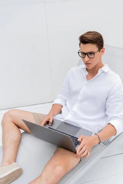 Thoughtful man in white shirt and eyeglasses using laptop in deck chair outdoors — Stock Photo