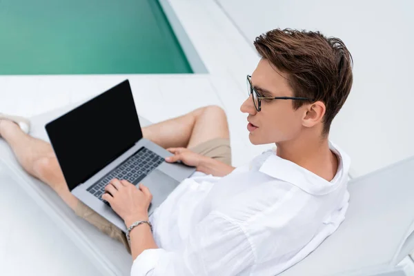 High angle view of man in eyeglasses sitting in deck chair near pool and using laptop — стоковое фото