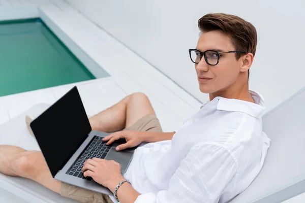 Man in eyeglasses looking at camera near laptop with blank screen in deck chair - foto de stock