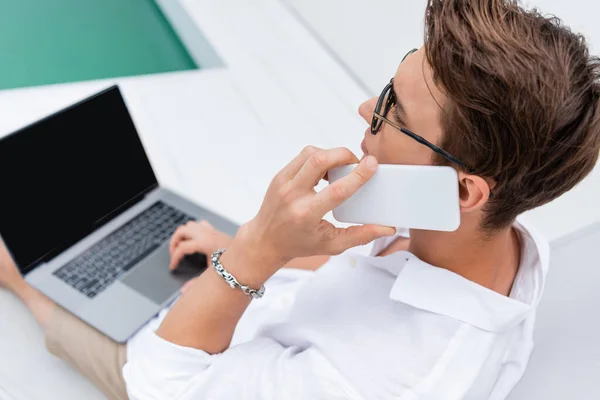 Высокоугольный вид человека, говорящего на смартфоне рядом с ноутбуком с экраном у бассейна — стоковое фото