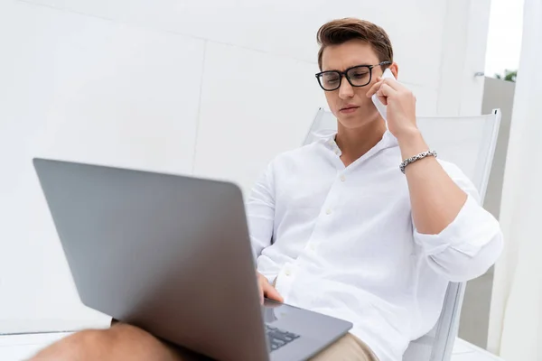 Serious man in eyeglasses looking at laptop and talking on cellphone in deck chair — Stock Photo