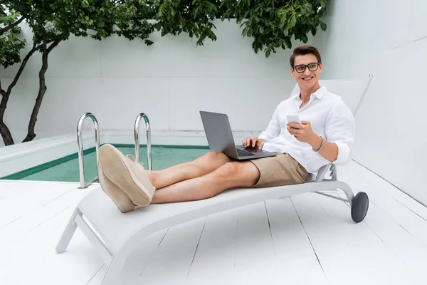 Full length of joyful man with gadgets looking away while sitting in deck chair near pool — Photo de stock