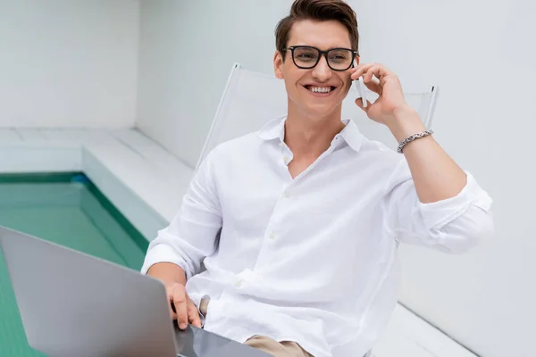 Cheerful man talking on smartphone near laptop and pool while looking away — стоковое фото