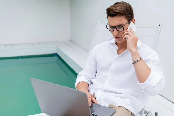 Freelancer in white shirt and eyeglasses using laptop while talking on smartphone near pool — стоковое фото