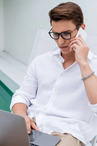 Focused freelancer in white shirt and eyeglasses using laptop and talking on cellphone outdoors - foto de stock