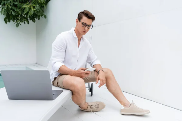 Full length of man in sneakers sitting at poolside near laptop and messaging on mobile phone — Fotografia de Stock