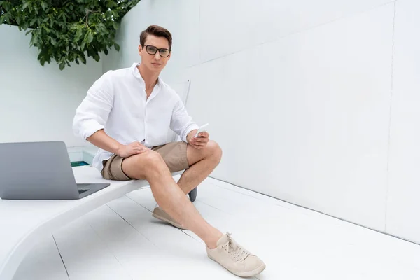 Full length of man in summer clothes sitting in deck chair with smartphone near laptop — Stockfoto