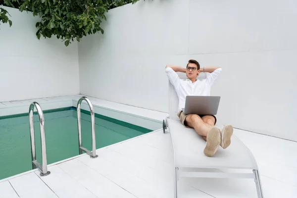 Full length of man with laptop relaxing in deck chair near pool — Stock Photo