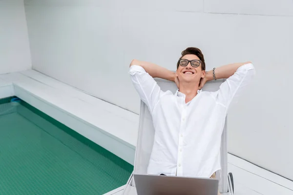 Happy man with laptop relaxing in deck chair near pool — стоковое фото