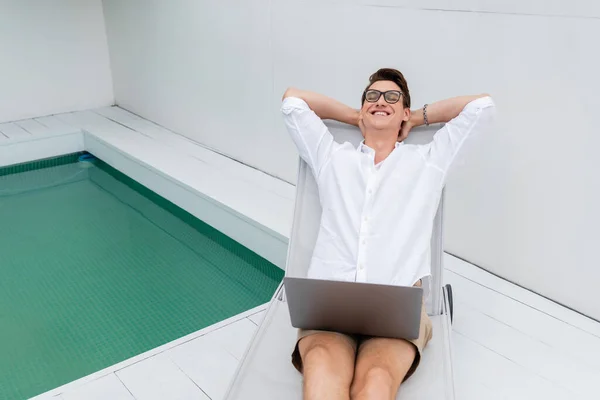 Happy man with closed eyes resting in deck chair with laptop near pool — Stockfoto