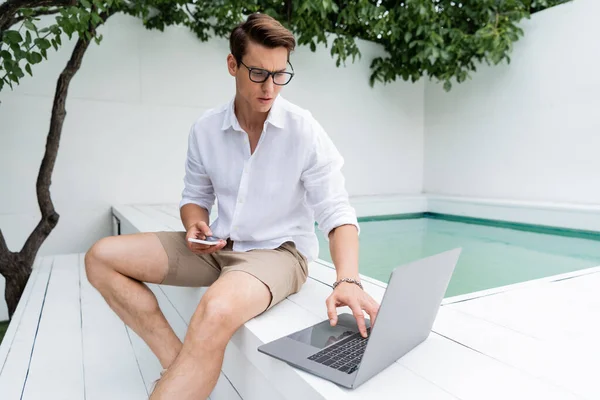 Вдумчивый человек в шортах сидит возле бассейна с мобильным телефоном и с помощью ноутбука — стоковое фото