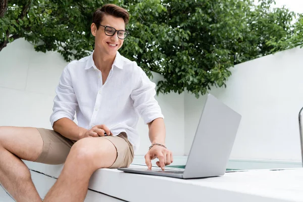 Glücklicher Mann in Sommerkleidung am Pool sitzend und auf Laptop schauend — Stockfoto