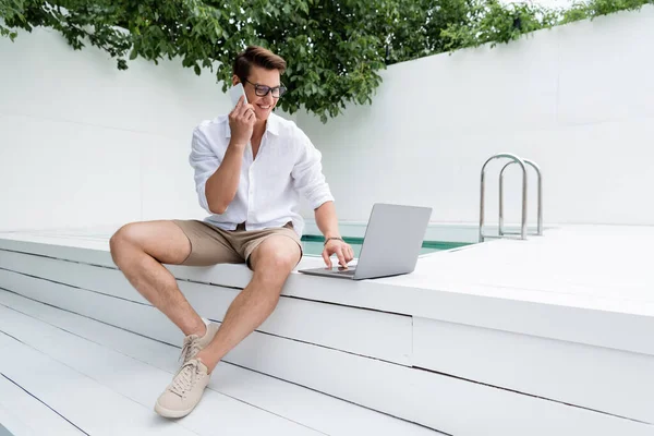 Piena lunghezza di freelance parlando sul cellulare durante l'utilizzo di laptop a bordo piscina — Foto stock